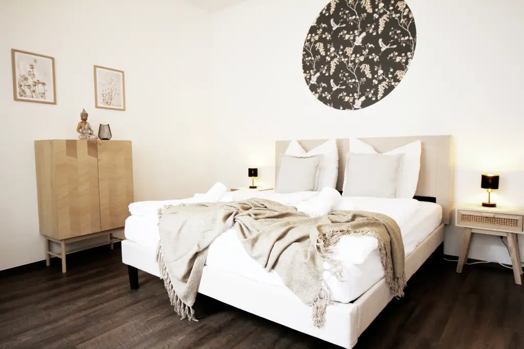 Schlafzimmer mit weißem Bett, Holzsideboard und rundem Blumenbild