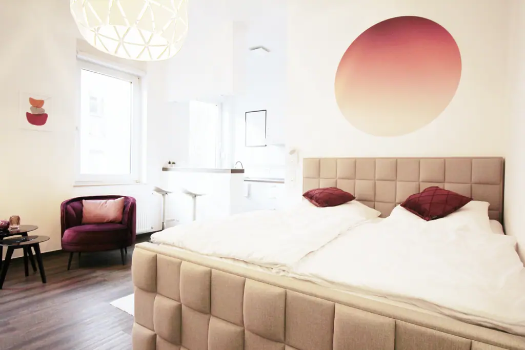 cozy 1.0: beiges Polsterbett mit rotem Wandbild und Sessel