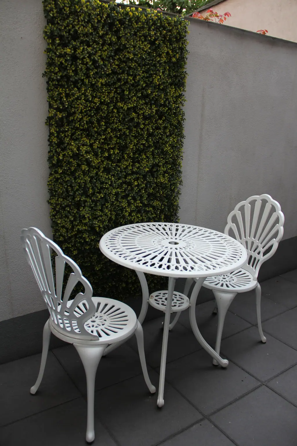 Kleine Terrasse mit weißem Bistrotisch und Stühlen vor einer Heckenwand